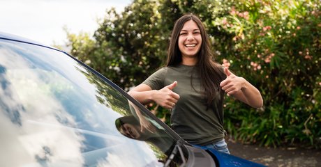 young girl holding her thumbs up in front of a blue vehicle